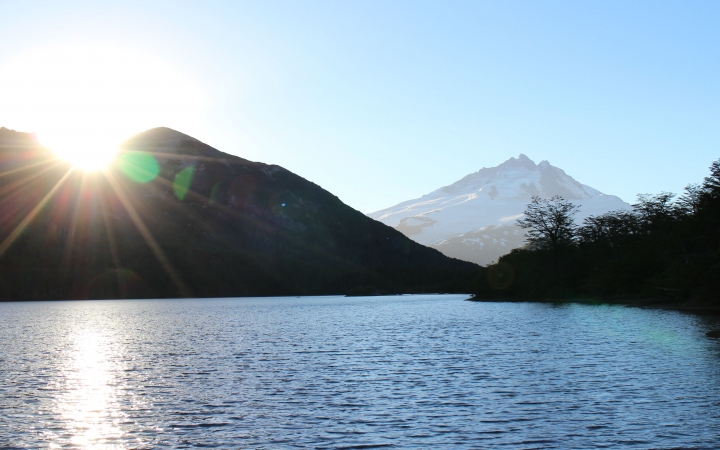 A blue body of water is framed by mountains and trees. In the background, there is a snow-covered mountain. 